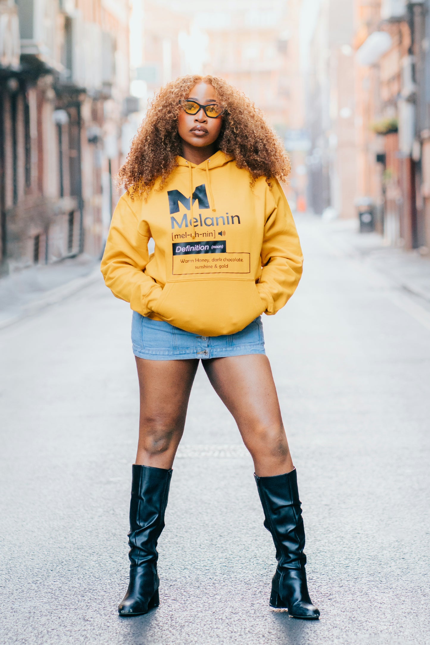 Women’s mustard yellow Melanin definition hoodie with front graphic print, styled with denim skirt and black boots.