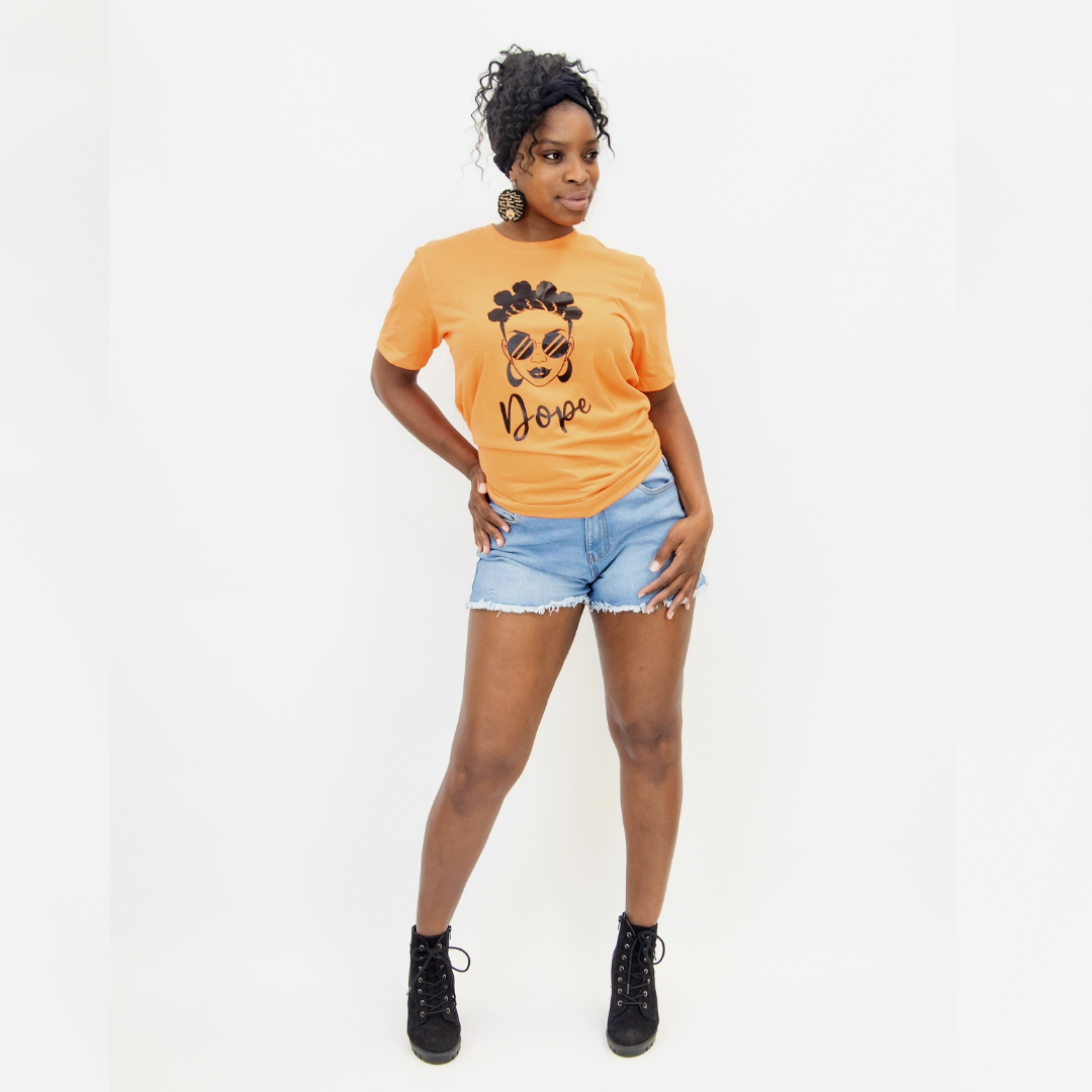 Full body view of woman in Afro Armour orange Dope and Bantu knots graphic t-shirt, denim shorts and black boots.